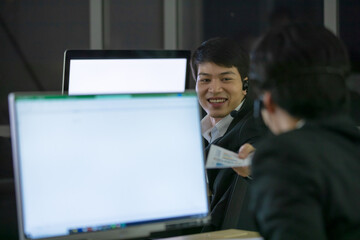 Asian smile man wearing headphone headset and working with happy face and sevice mind in call center in front of computer screen in office at night. Overtime, late work and 24 hours service concept