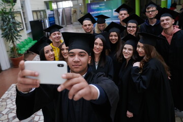 Obraz premium group of happy international students in mortar boards and bachelor gowns with diplomas taking selfie by smartphone