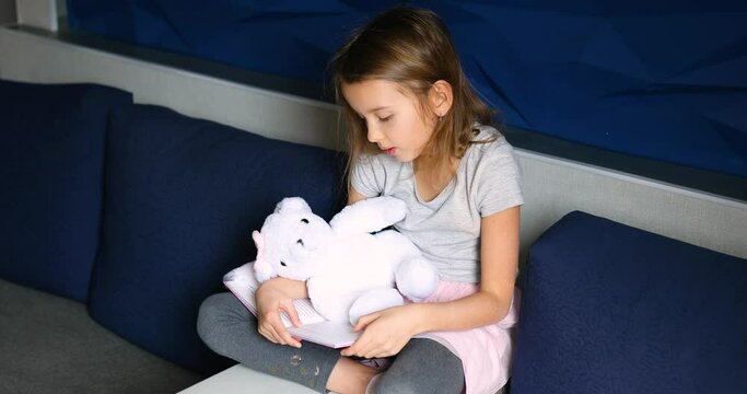 Cute little girl eading a book with stuffed teddy bunny toy and smiling sitting on the sofa in the living room. Stay at home during coronavirus covid-19 pandemic