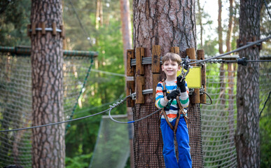 Young cute child boy in summer clothing, safety harness and helmet attached with carbine to cable moves slowly along rope way on green trees sunny bokeh background. Sport, game, leisure concept