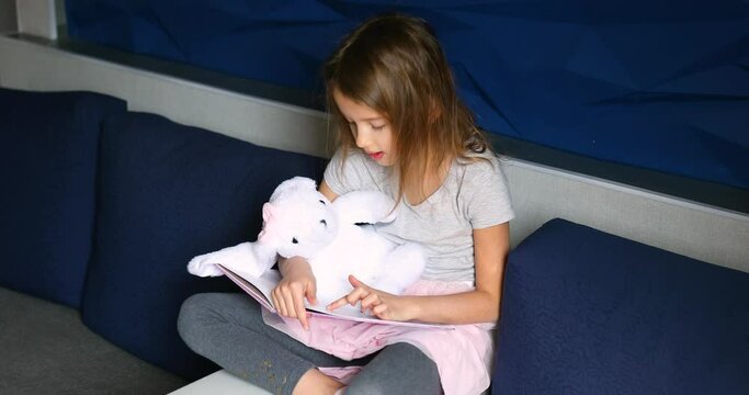 Cute little girl eading a book with stuffed teddy bunny toy and smiling sitting on the sofa in the living room. Stay at home during coronavirus covid-19 pandemic