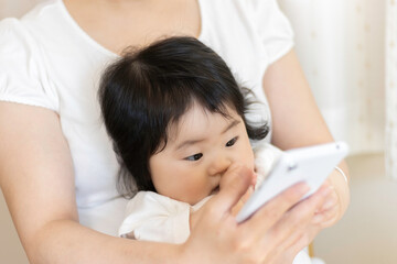 赤ちゃんを膝に座らせて、スマートフォンを使う女性。赤ちゃん（0歳、生後8か月、日本人、女の子）