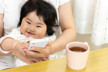赤ちゃんを膝に座らせて、スマートフォンを使う女性。赤ちゃん（0歳、生後8か月、日本人、女の子）