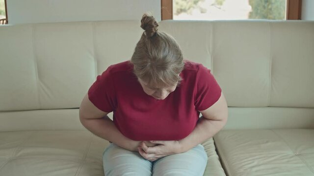 Portrait of a caucasian woman with blonde hair sitting on sofa having strong stomach ache hands on abdomen