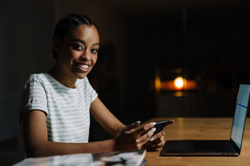 Black smiling woman using mobile phone while working with laptop