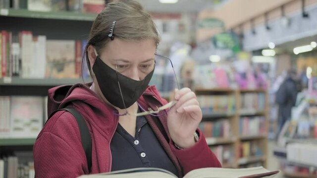 Serious woman wearing glasses and a mask against the virus reads a book in the library. Then she closes eyes and takes off his glasses. The concept of visual impairment, diseases and eye fatigue