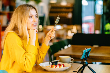 Dreamy female blogger eat cheese pancakes with jam while sitting at the restaurant table using smartphone. woman thinking about something, pensive female breakfast in coffee shop.