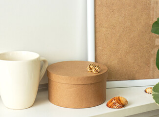 Still life. Cup of coffee, blank beige poster frame, craft round box and beautiful gold earrings on the white wall. Home decor