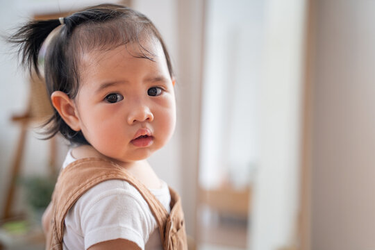 portrait of asian baby infant in brown standing turn back looking at camera in living room at home on day time. baby response when calling.