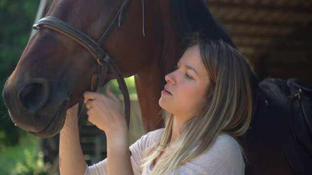 Close up of young woman preparing her horse, putting bridle and harness. Woman fixing mane from harness in a riding school