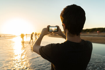 Young man on the beach taking a picture of the sun at sunset.