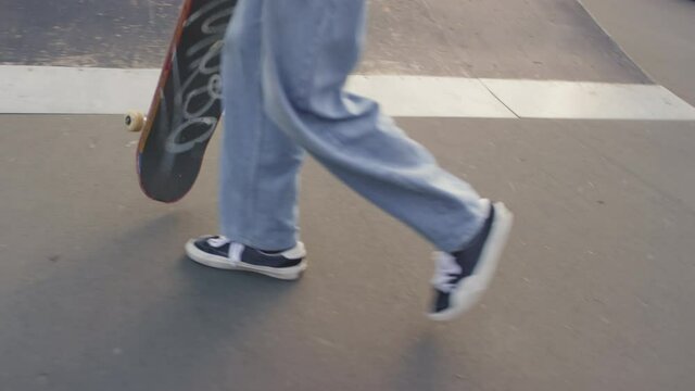 Handheld low-section shot of unrecognizable teenager in jeans carrying skateboard and walking in skatepark
