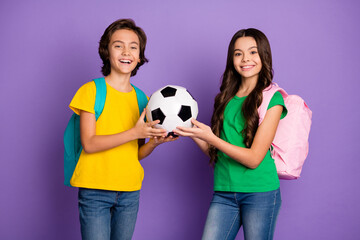 Photo of two cheerful boy and girl hold football wear backpack school isolated on violet color...