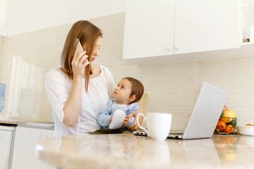 Beautiful business mom is talking on the mobile phone and taking notes while spending time with her cute baby boy at kitchen home office. Working mother concept,Laptop on table