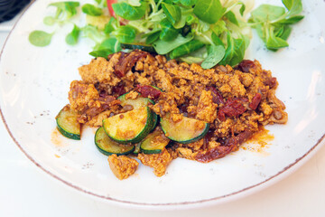 Vegan breakfast prepared with scrambled tofu and eggs, cooked with dried tomatoes and zucchini served with fresh lettuce and cucumber in white ceramic vintage plate