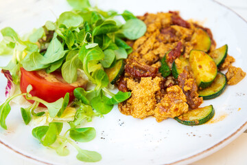 Vegan breakfast prepared with scrambled tofu and eggs, cooked with dried tomatoes and zucchini served with fresh lettuce and cucumber in white ceramic vintage plate