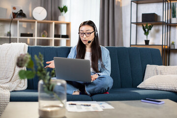 Confident young woman in headset sitting on couch and having video conference on laptop. Female freelancer leading working meeting online while staying at home.