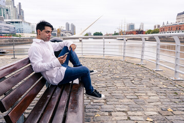 Fototapeta na wymiar Young Latino man sitting on a wooden plaza bench looking at the old port of the city. Urban scene.