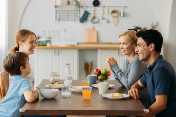 Obraz na płótnie Canvas Smiling caucasian family of man and woman sitting together and listening to small and cute son with daughter while eating healthy breakfast at home