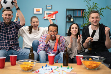 Happy mixed-races friends resting in the evening at home with beer and cheering up for a goal on...