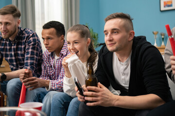 A young students keeps fingers crossed for his football team. A group of friends of different nationalities watch the game while sitting on the living room couch at home.