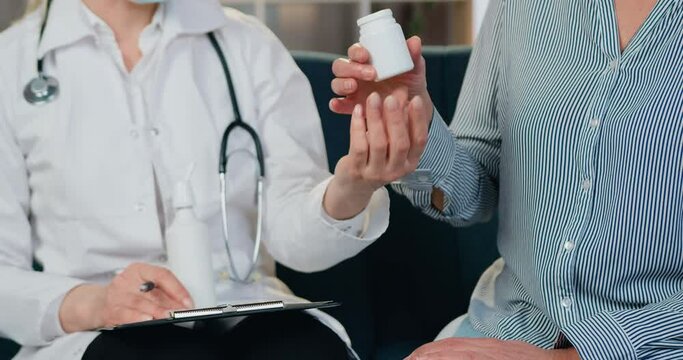 Attractive experienced professional female doctor in facial mask and white gawn explaining how to take tablets or vitamins to her female patient during home visit