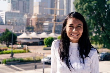 Mid-shot portrait of a black-haired Latina woman smiling, with the city in the background. - obrazy, fototapety, plakaty