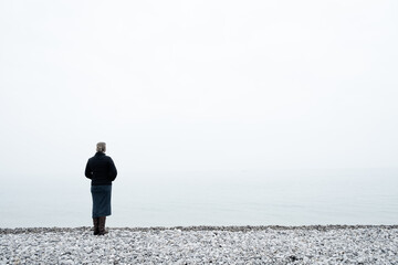 Woman wearing warm clothes and a purse looking out over a foggy sea from a pebble beach on the shore
