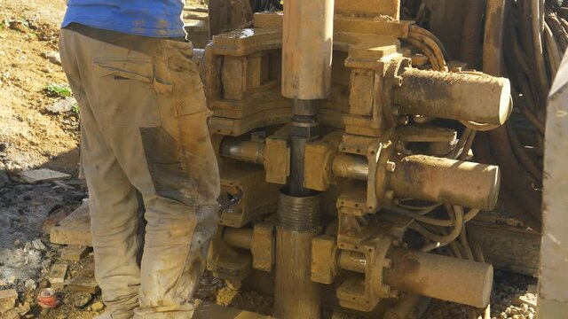 CLOSE UP: Unrecognizable male contractor stands next to a drill boring a hole in the ground near a construction site. Worker oversees the boring process during modern geothermal pump installation.