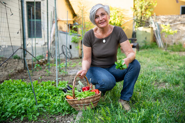 Fototapeta na wymiar Portrait of an adult woman with the basket with the vegetables harvested in the urban garden in her field near the house - Concept of sustainability and self-maintenance