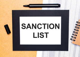 On a wooden table, there is a black open marker, a brown notepad and a sheet of paper in a black frame with the text SANCTION LIST. View from above.