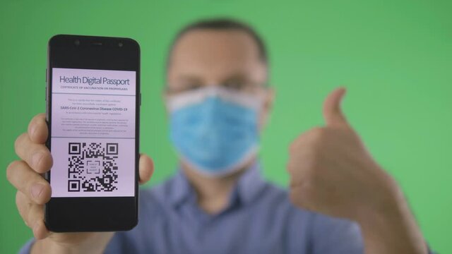 Young man with face mask shows smartphone for scanning the digital health certificate, thumb up OK sign to encourage others to use it, front view isolated on green screen chroma key