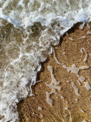 Close-up of the sea coast, clear sea water with shallow waves and a narrow strip of white foam with bubbles washes the shore with a smooth brown sandy surface. High quality photo