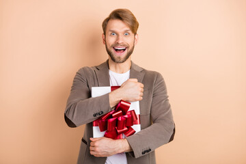 Photo of impressed funny young guy wear jacket smiling hugging gift box open mouth isolated beige...