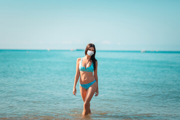 Fototapeta na wymiar Summer. A young pretty woman wearing a protective mask poses standing at the ocean. The concept of summer holidays during the coronavirus pandemic