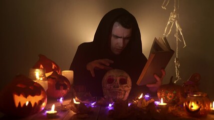 A spooky wizard is reading a magic book and conjuring over the skull on the black background. A young caucasian man in a gothic cloak is laughing ominously surrounded by Halloween decorations and fume
