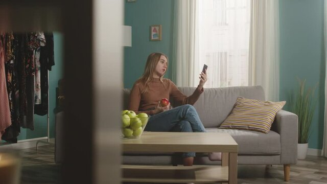 Woman in blue jeans and brown sweater sits on beige sofa on window background, bites red apple and talks on video call. On coffee table is glass jar with green apples. Camera dollies horizontally