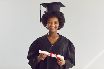 Happy smiling young african american smart woman student in academic hat and gown holding diploma graduation degree certificate scroll looking at camera standing against gray studio background.