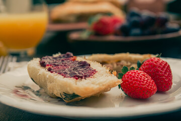 Slice of bread with strawberry jam and strawberries on a white plate. Breakfast unfocused at the background.