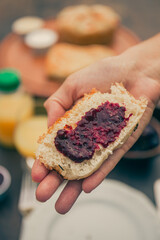 Hand holding a slice of bread with strawberry jam on the foreground. Breakfast unfocused at the background.