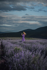 a blonde woman stands in a purple dress in the mountains on a lavender field in the rays of sunlight