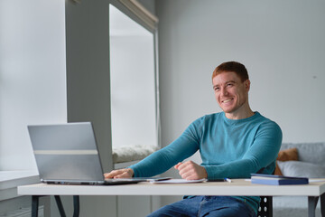 man is at home at his desk uses a laptop smiling and showing a like to his colleague via video communication.