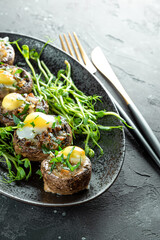 Baked champignons with quail eggs and microgreen garnish on a black plate close up vertical photo