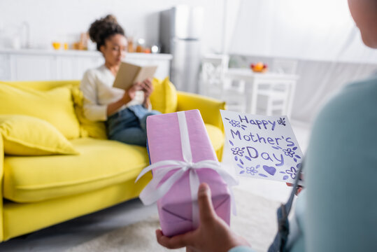 kid holding happy mothers day card near african american woman reading book on sofa, blurred background