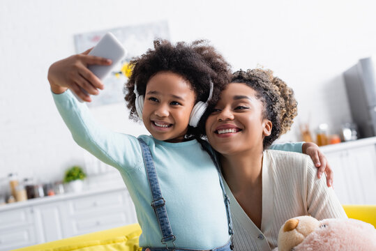 cheerful african american girl in headphones taking selfie on smartphone with happy mother