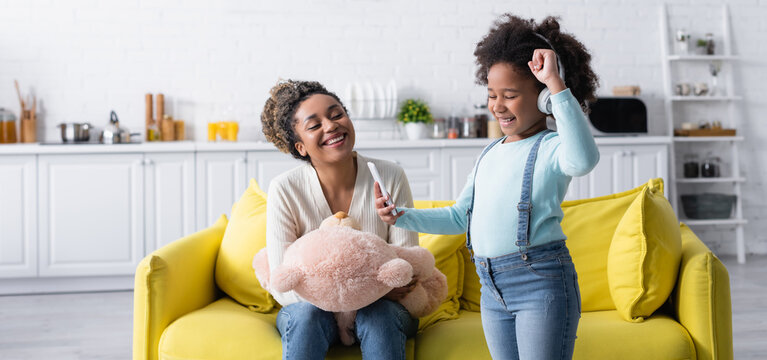 excited african american child in headphones showing win gesture near mother with teddy bear, banner