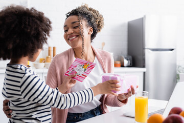 african american child presenting gift and mothers day greeting card to happy mom in kitchen, blurred foreground