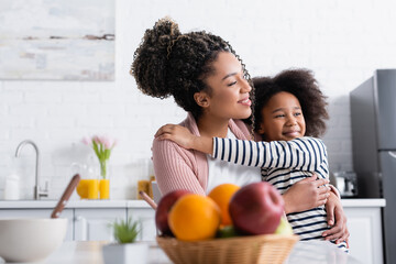 cheerful african american woman and child embracing while looking away near fruits on blurred foreground