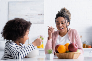 cheerful african american woman looking at daughter while holding spoon with corn flakes, blurred foreground