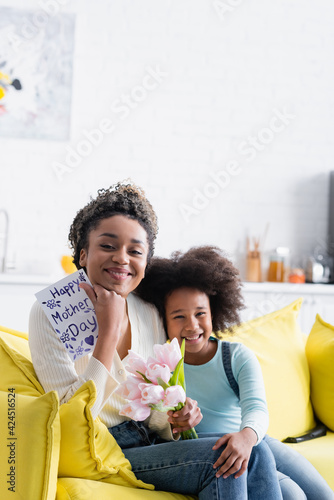 joyful african american girl sitting near happy mom holding tulips and happy mothers day card